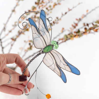 Hand holding a stained glass dragonfly suncatcher with blue and white wings, green body, and delicate details.