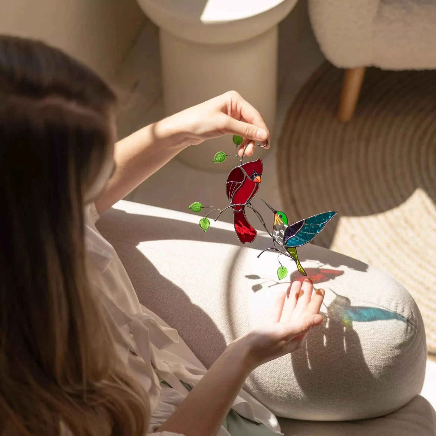 Person holding a colorful stained glass cardinal with hummingbird ornament in a softly lit room.