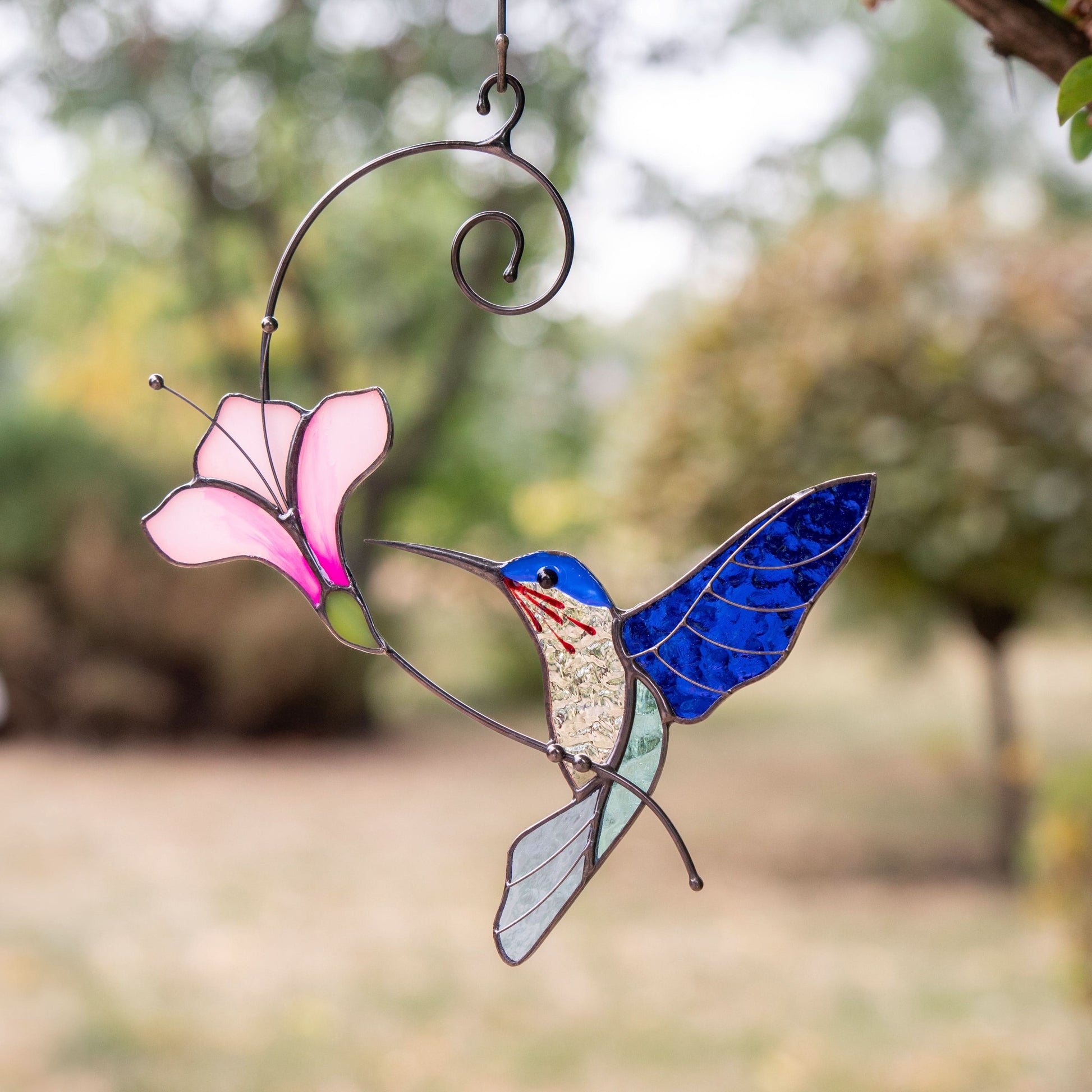 Blue stained glass hummingbird with pink flower hanging outside among trees and soft greenery