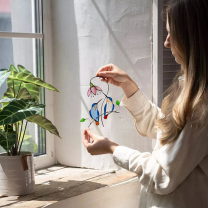 Person holding a colorful stained glass pair of hummingbird window hanging near a window with a plant on a windowsill.
