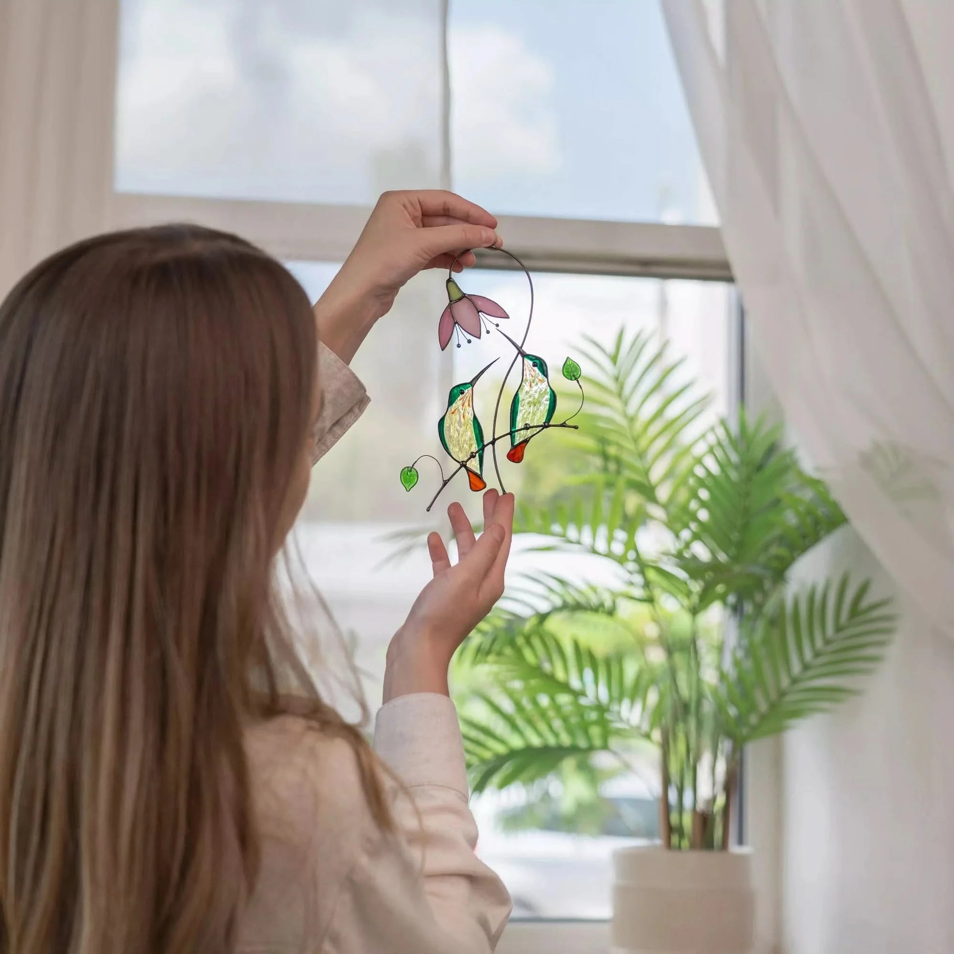 Person holding a stained glass pair of hummingbirds decoration in front of a window with plants