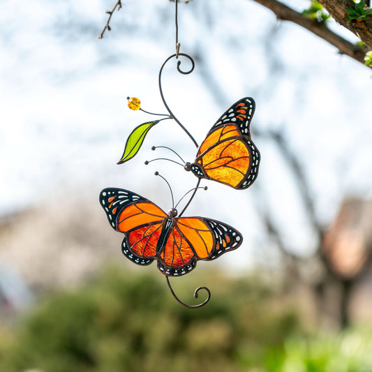 Two stained glass monarch butterflies displayed outdoors among tree branches, glowing brightly in natural sunlight.