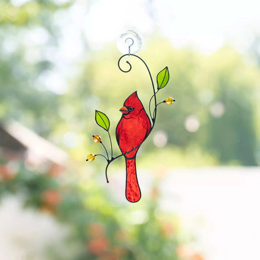 Stained glass cardinal bird hanging on a branch with leaves and flowers against a blurred green outdoor background.