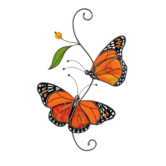 Two stained glass monarch butterflies in vibrant orange shades with a green leaf accent, hanging near a white vase with yellow flowers.