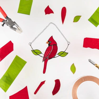 Cardinal suncatcher in the center surrounded by red and green stained glass pieces and tools