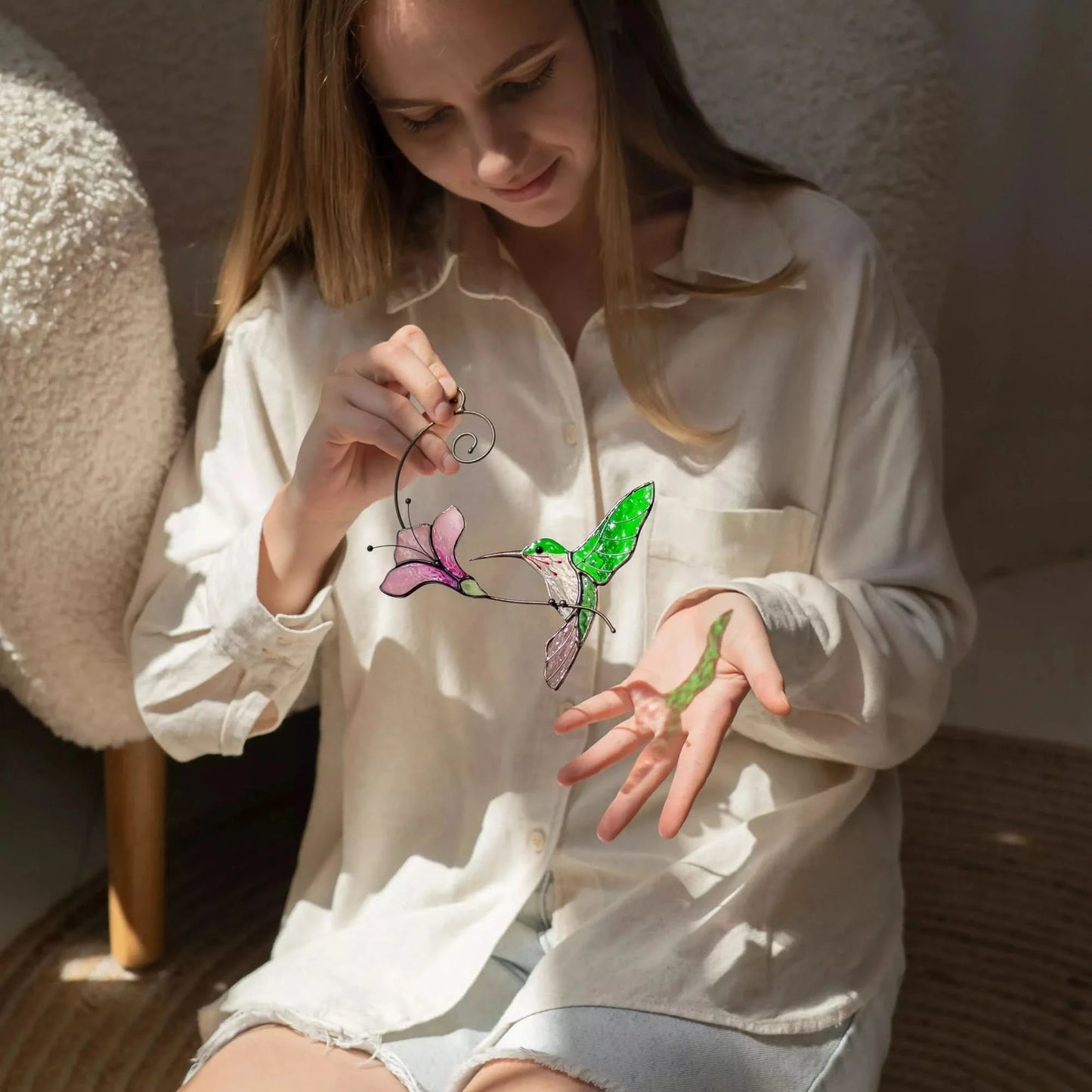 Woman holding colorful hummingbird suncatcher, that casts soft colorful shadow, in a softly lit room
