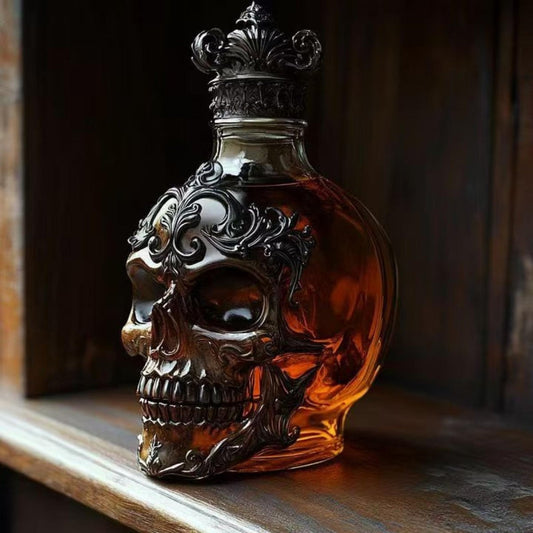 Side profile of the Kouvr ornate skull whiskey decanter filled with amber liquid, showing detailed metal filigree on a dark wood shelf.